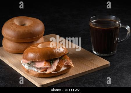 Ein Sandwich mit Lachs und Dill liegt auf einem Holzbrett neben einer Tasse Kaffee. Das Sandwich wird auf einem Holzbrett platziert und der Kaffee ist in einem gla Stockfoto