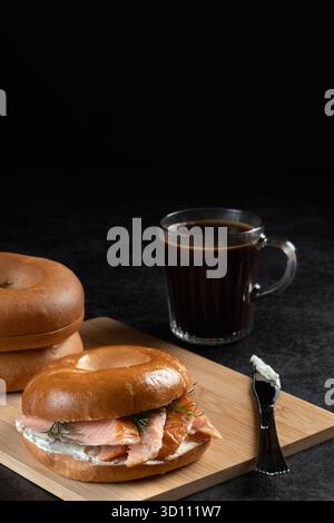 Ein Sandwich und eine Tasse Kaffee stehen auf einem Holztisch. Das Sandwich ist ein Bagel mit Lachs und Dill. Der Kaffee ist schwarz und in einem Glas. Szene ist Casua Stockfoto