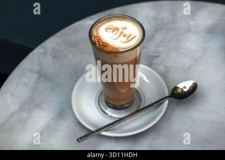 Nahaufnahme von Latte Coffee in einer transparenten Tasse mit der Aufschrift „Enjoy“ auf dem Milchschaum Stockfoto