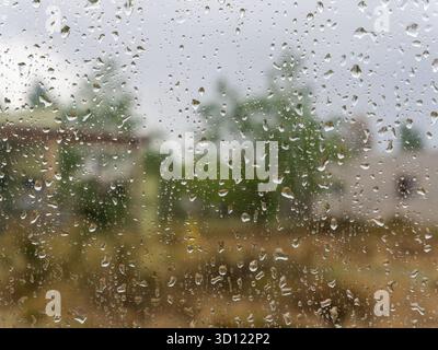 Wassertropfen, die ein Glasfenster bedecken, zeigen einen weichen Hintergrund einer Wohnszene mit natürlichen Elementen, während es noch regnet Stockfoto