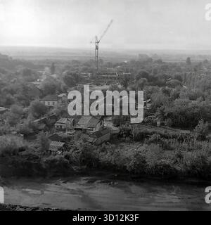 Ein Panoramablick aus einem hohen Winkel auf das Viertel Khimik in Sloviansk, 1970er-1980er Jahre Dieses archivierte Schwarzweißfoto aus der UdSSR, der ukrainischen SSR, zeigt den Rand der Stadtentwicklung. Ein großer Bereich flacher Privathäuser („chastnyy Sektor“) mit Gärten ist hervorzuheben. Im Hintergrund weist ein hoher Turmkran auf den Bau neuer mehrstöckiger sowjetischer Wohnblöcke („Panelka“) hin, was auf die Erweiterung des „Schlafviertels“ hindeutet. Damit wird der typische Landschaftskontrast erfasst. Ein wertvolles Dokument über das friedliche Leben im Donbass vor dem Krieg. Stockfoto