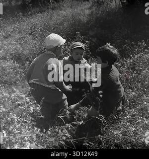 Drei sowjetische Kinder, ein älterer Junge, ein Mädchen und ein Kleinkind, spielen zusammen im Freien in Hochrasen in Sloviansk, 1970er Jahre Dieses Archivfoto aus der UdSSR (Ukrainische SSR) zeigt eine offene Szene der Kindheit während der Breschnew-Ära. Die Kinder tragen typische Kleidung der 1970er Jahre. Das Mädchen hat kurze Haare. Sie sind in ihrem Spiel in einer natürlichen Umgebung vertieft. Dieses nostalgische Bild erinnert an die einfachen Freuden der sowjetischen Kindheit. Ein wertvolles Dokument über das friedliche Leben im Donbass vor dem Krieg. Stockfoto