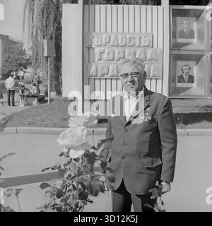 Ein Porträt eines dekorierten älteren sowjetischen Mannes, wahrscheinlich ein Veteran der Arbeit, der stolz in Sloviansk steht, 1970er Jahre Dieses Archivfoto aus der UdSSR, der ukrainischen SSR, zeigt den Mann mit Brille und einer Anzugsjacke, die mit sowjetischen Medaillen und Orden geschmückt ist. Er posiert vor dem Ehrenamt der Stadt („Doska Pocheta“), das Porträts vorbildlicher Bürger unter dem Titel „Stolz der Stadt und des Bezirks“ zeigt. Dieses Bild fängt das Wesen der sowjetischen öffentlichen Anerkennung und des Respekts vor Leistungen ein. Ein wertvolles Dokument über das friedliche Leben im Donbass vor dem Krieg. Stockfoto
