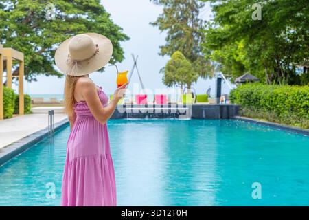 Frau in lila Kleid am Pool mit Cocktail, tropisches Resort, Stockfoto