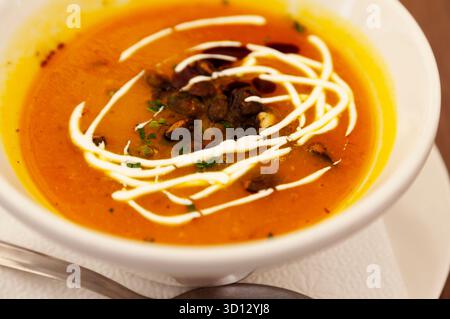 Eine Schüssel mit leuchtender Orangen-Kürbissuppe, garniert mit einem künstlerischen weißen Creme-Strudel und dunklen Kräutern oder Gewürzen. Die Suppe wird in einer weißen Keramikschleife serviert Stockfoto