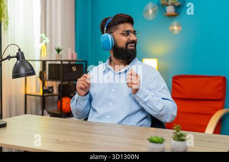 Indischer Mann sitzt zu Hause glücklicher Ausdruck, der Kopfhörer trägt, schwingt sanft tanzen und genießt ruhige, sanfte Musik. Arabian an der Rezeption hält Rhythmus Lächeln fühlt sich friedliche Stimmung und genießt Melodiemomente tief. Stockfoto