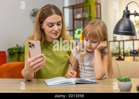 Mutter und Tochter sehen zu Hause Online-Unterricht auf dem Smartphone Schreiben Sie Notizen in Notizbuch Folgen Sie Aufgaben. Frau und Mädchen am Tisch überprüfen Übung markieren Antworten Fehler beheben Regeln Praxis Fokus Tempo gut lernen Stockfoto
