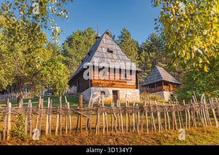 Traditionelles serbisches ländliches Blockhaus mit einem rustikalen Zaun, eingebettet in einen grünen Wald unter einem klaren blauen Himmel Stockfoto