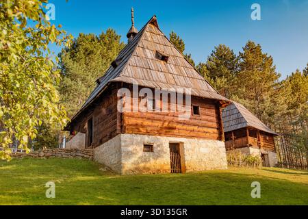 Traditionelles serbisches ländliches Blockhaus mit einem rustikalen Zaun, eingebettet in einen grünen Wald unter einem klaren blauen Himmel Stockfoto