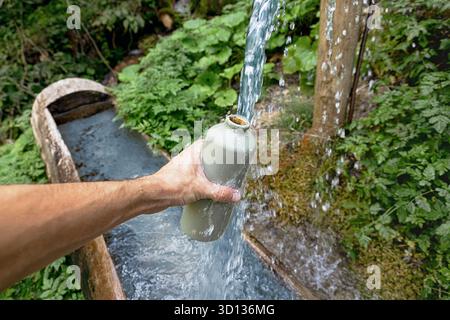 Hand hält eine wiederverwendbare Wasserflasche und füllt sie mit frischem, fließendem Wasser aus einem rustikalen Holzauslauf in einer natürlichen, grünen Waldumgebung Stockfoto