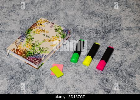 Schreibwaren wie ein Notizbuch mit Blumenmotiv, Haftnotizen und leuchtende Textmarker sind auf einer strukturierten Oberfläche angeordnet und verleihen dem Arbeitsplatz ein angenehmes Ambiente Stockfoto