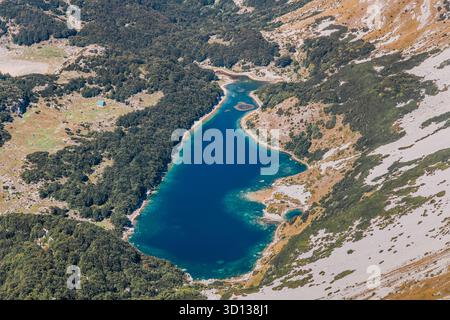 Aus der Vogelperspektive entdecken Sie einen ruhigen Bergsee, umgeben von dichtem Wald und zerklüfteten Gipfeln, mit einer kleinen Hütte darunter Stockfoto