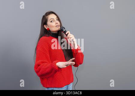 Junge, emotionale Brünette mit einem Mikrofon in einem roten Pullover singt Karaoke oder sagt eine Rede, isoliert auf Grau Stockfoto