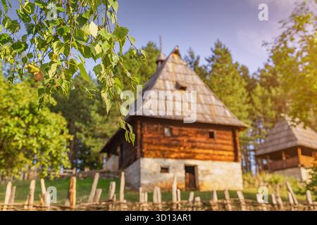 Verschwommenes traditionelles serbisches Landhaus mit einem rustikalen Zaun, eingebettet in einen grünen Wald unter einem klaren blauen Himmel Stockfoto