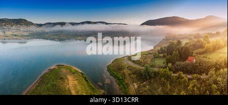 Friedliche Morgenszene mit einem ruhigen See, weichem Nebel, der die umliegenden Hügel bedeckt, und Sonnenlicht beleuchtet die Landschaft Stockfoto