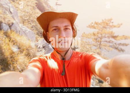 Ein Mann lächelt in die Kamera und macht Selfie in orangefarbenem Hemd und Hut, während er auf einem sonnendurchfluteten Bergpfad wandert Stockfoto