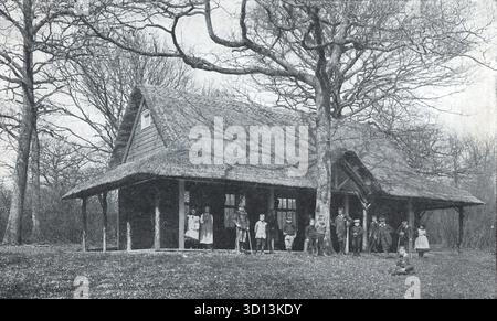 Schwarzweiß-Fotografie aus der Zeit um 1908, herausgegeben von der Queen Alexandra League. Stellt die Forest School am Lord Mayor Treloar Hospital in Alton, Hampshire dar. Ein rustikales Strohdach-Gebäude steht inmitten von blattlosen Bäumen, in dem Kinder und Erwachsene in alten Kleidern draußen posieren. Die Szene spiegelt die Bildung im Freien aus der edwardianischen Zeit und die Erholungspflege in einem bewaldeten institutionellen Umfeld wider. Von einer Originalpostkarte. Stockfoto