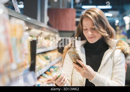 Lächelnde blonde Frau untersucht ein Glas Joghurt im Lebensmittelgeschäft, vergleicht seine Zutaten und Haltbarkeit auf ihrem Telefon, um gesund zu machen Stockfoto