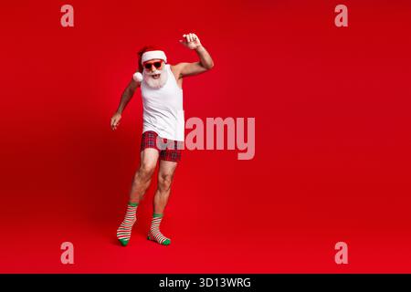 Energiegeladener Weihnachtsmann mit Hut weißem Tanktop karierte Shorts, der in einem roten Studio für festliche Feiertage springt – Stockfoto Stockfoto