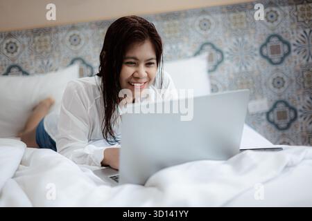 Asiatische Frau, die auf dem Bett liegt, mit einem Laptop für Fernarbeit, Online-Lernen und digitale Kommunikation im gemütlichen Schlafzimmer, Konzept des Wohn-Lifestyle und Technologie Stockfoto