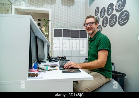 Lächelnder Physiotherapeut sitzt auf einem Trainingsball, während er an einem Computerpult arbeitet Stockfoto
