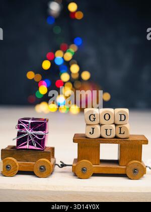 Gott Jul, Scaninavian Merry Christmas mit einem kleinen hölzernen Zug Set mit Geschenkbox auf verschwommenen Weihnachtslichtern, saisonaler oder weihnachtlicher Hintergrund Stockfoto