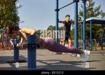 Junge Erwachsene kaukasische Frau, die Liegestütze auf parallelen Bars im Freien macht, während junger Erwachsene kaukasische Mann im Hintergrund mit ausgestreckten Armen in der Nähe von Outdoor-Fitnessgeräten steht Stockfoto