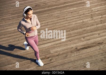 Junge Erwachsene kaukasische Frau, die draußen auf hölzernen Boardwalk joggt, Kopfhörer trägt, mit fokussiertem Ausdruck nach vorne blickt, Fitness-Aktivitäten nachgeht, allein trainiert Stockfoto
