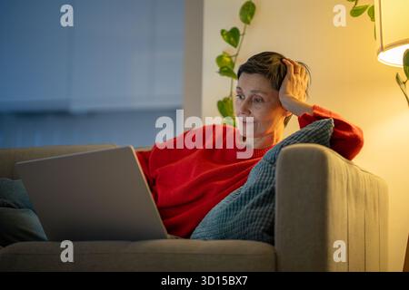Entspannte Frauen liegen auf dem Sofa mit Laptop. Reife Frau, die fernsehserien auf dem Streaming-Dienst ansieht Stockfoto