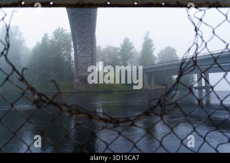 Historische Heinola-Eisenbahnbrücke durch eine Lücke in einem Maschendrahtzaun an einem nebeligen Morgen. Stockfoto