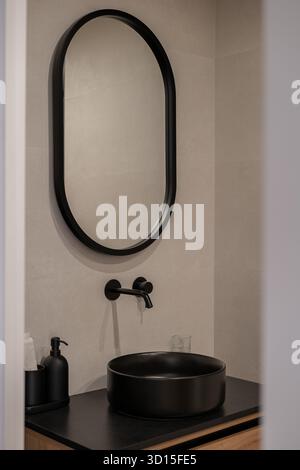 Das moderne Badezimmer verfügt über ein rundes schwarzes Waschbecken, einen eleganten schwarzen Wasserhahn und einen großen ovalen Spiegel. Die Wände sind hell, was einen minimalistischen Stil schafft Stockfoto