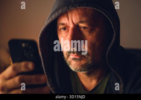 Mann trägt schwarzen Hoodie mit Smartphone in einem dunklen Raum mit warmer dezenter Beleuchtung und stimmungsvoller Atmosphäre. Selektiver Fokus. Stockfoto