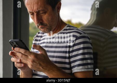 Reifer Mann in gestreiftem Matrosenhemd, der auf dem Balkon steht und auf dem Smartphone SMS schreibt, reflektiert in Fensterglas, symbolisiert moderne Kommunikation und Soli Stockfoto
