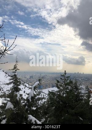 Von Tochal aus überblicken schneebedeckte Nadelbäume die Skyline von Teheran, während sich das Licht durch übereinander liegende Wolken bricht. Stockfoto