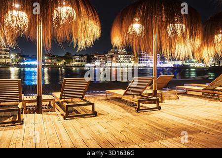 Leere hölzerne Sonnenliegen unter Strohschirmen auf einer Terrasse am Wasser bei Nacht mit Lichtern, die auf dem Wasser c'an pastilla mallorca reflektieren Stockfoto