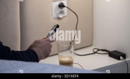 Nahaufnahme eines Mannes, der ein Smartphone neben einem Bett hält, mit einem Glas Kaffee und Ladekabeln auf einem weißen Nachttisch. Die gemütliche Abendszene illustriert Stockfoto