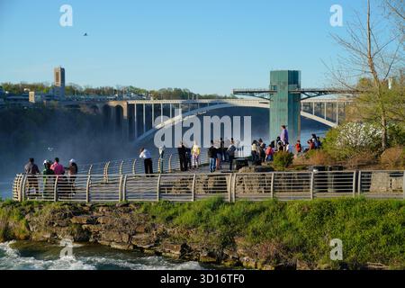 Touristen versammeln sich auf dem Aussichtsgebiet im Niagara Falls State Park, wo die Regenbogenbrücke und der Nebel von den Wasserfällen im Hintergrund sichtbar sind Stockfoto
