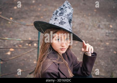 Ein Teenager mit Hexenhut schaut in die Kamera, während er die Krempe anpasst, um ein selbstbewusstes Halloween-Porträt zu schaffen Stockfoto