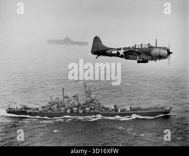 VOR DEN GILBERT-INSELN, PAZIFIK - 12. November 1943 - Eine US-Navy Douglas SBD-5 Dauntless der Bombenstaffel VB-16 fliegt eine U-Boot-Patrouille tief über dem Schlachtschiff USS Washington (BB-56) auf dem Weg zur Invasion der Gilbert-Inseln während des Zweiten Weltkriegs. Das Schiff im Hintergrund ist USS Lexington (CV-16), der Heimträger des Flugzeugs - Foto: Geopix/US Navy Stockfoto