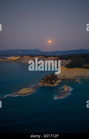 Aus der Vogelperspektive auf einen Vollmond, der über den atemberaubenden Küstenklippen von Kap Drastis, Korfu, aufsteigt. Das Ionische Meer leuchtet in der Abenddämmerung mit den Lichtern der Stadt in der di Stockfoto