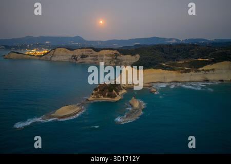 Majestätische Aussicht auf den Vollmond, der über den einzigartigen weißen Klippenformationen des Kap Drastis auf Korfu, Griechenland, aufsteigt, mit Lichtern der Stadt Stockfoto