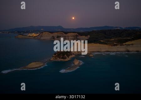 Dramatische Luftaufnahme des Vollmondes über Cape Drastis, Korfu. Das dunkle Ionische Meer und die weißen Klippen werden vom Mond und den fernen Lichtern der Stadt beleuchtet Stockfoto