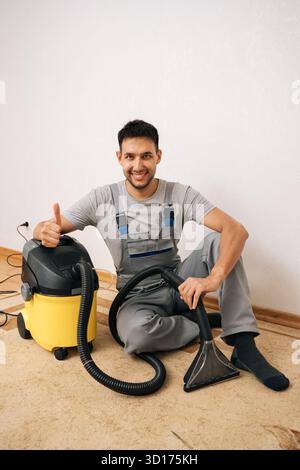 Freundlicher Reinigungsservice-Angestellter, der insgesamt die Daumen nach oben zeigt und auf dem Boden sitzt, mit einem Waschstaubsauger, bereit für einen gründlichen Boden Stockfoto