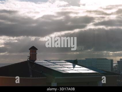 Bedeckter Himmel mit Sonnenstrahlen, die durch Wolken brechen und sich auf Sonnenkollektoren auf dem Dach spiegeln. Dramatische Lichter und Kontraste. Stockfoto