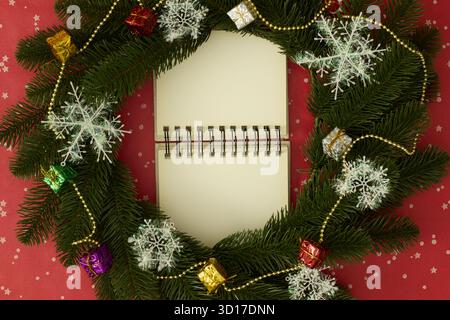 Offenes Notizbuch, eingerahmt von einem Weihnachtskranz mit Schneeflocken, Geschenkboxen und Goldperlen auf rotem Hintergrund Stockfoto