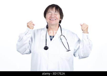 Glückliche Ärztin im Laborkittel und Stethoskop hebt mit geschlossenen Augen die Fäuste im Sieg, lächelnd zur Feier einer erfolgreichen Behandlung Stockfoto