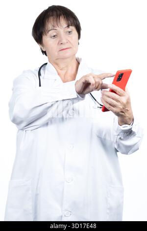 Erfahrene Ärztin, die einen Labormantel und ein Stethoskop trägt, ein rotes Smartphone ansieht und darauf scrollt, während sie virtuelle Gesundheitsdienste anbietet Stockfoto