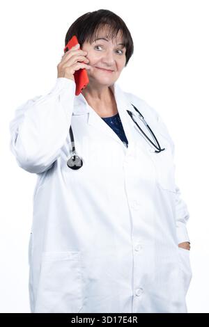 Erfahrene Ärztin mit Laborkittel und Stethoskop, die lächelnd auf einem roten Smartphone spricht und Telekonsultation repräsentiert Stockfoto