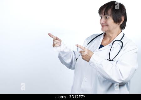 Lächelnde Seniorenärztin trägt einen weißen Labormantel und Stethoskop, zeigt mit beiden Händen, zeigt oder empfiehlt medizinische Informationen Stockfoto