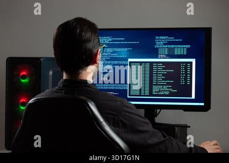 Nahaufnahme eines Mannes, der rückwärts einen Computermonitor mit einem blauen Programmiercode auf dem Bildschirm beobachtet Stockfoto